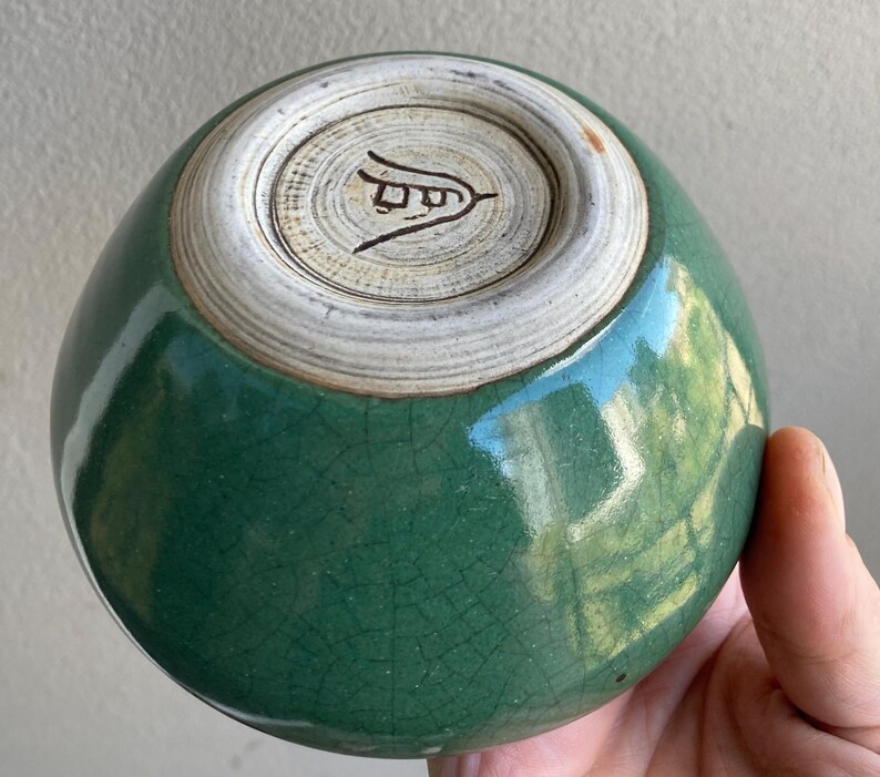 May include: A handcrafted, green ceramic bowl with a crackle glaze. The bowl's base is a lighter color with a stamped design. The bowl is held in a hand.