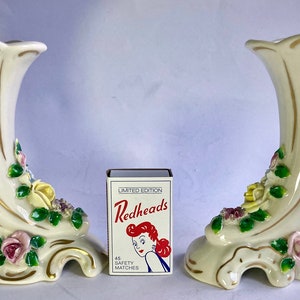 May include: Pair of white ceramic vases with gold trim and floral details. The vases are shaped like horns and have a scalloped rim. The flowers are pink, yellow, and green.