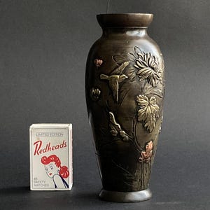 May include: Bronze-coloured vase with embossed floral and bird designs. The vase has a wide body and a narrow neck. A box of Redheads safety matches is next to the vase.