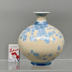 May include: A cream-coloured ceramic vase with a crystalline glaze featuring blue circular patterns. The vase has a rounded body and a flared neck. Next to the vase is a rectangular box of Redheads safety matches for scale.