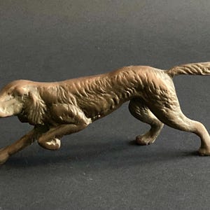 Può includere: Statuetta di cane color bronzo in posizione di corsa. Il cane ha un corpo lungo, una coda fluida e una texture di pelliccia dettagliata. La statuetta è su una superficie scura.