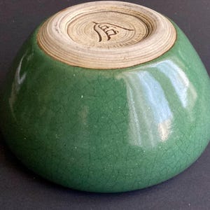 May include: A small, green ceramic bowl with a crackle glaze. The bowl has a rounded shape and a light-colored, unglazed base with a carved design. The exterior is a vibrant green color.