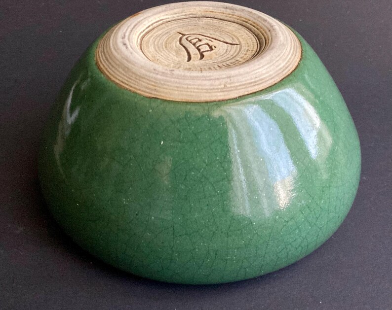 May include: A small, green ceramic bowl with a crackle glaze. The bowl has a rounded shape and a light-colored, unglazed base with a carved design. The exterior is a vibrant green color.