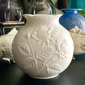 May include: A white ceramic vase with a round body and a ruffled rim. The vase features a raised relief design of roses, buds, and leaves. Other decorative objects are visible in the background, including a blue vase and a metal fish.