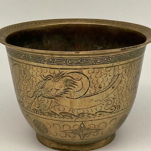 May include: A brass bowl featuring an etched dragon design encircling the exterior. The rim is decorated with a geometric pattern, while the lower section displays circular motifs and star-like shapes. The metal has a warm, aged golden tone.