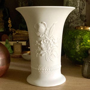 May include: A white ceramic vase with a flared top and a detailed floral relief design. The vase features a decorative band near the base and stands approximately 8 inches tall. The background includes other decorative items.