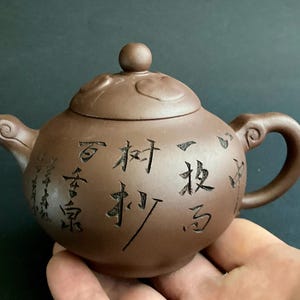 Puede incluir: Tetera de cerámica marrón con cuerpo redondeado, boquilla y asa. La tapa tiene un pequeño pomo y elementos decorativos. La tetera está inscrita con caracteres chinos negros. Una mano sostiene la tetera.