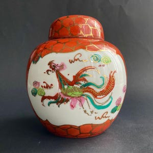 May include: A ceramic jar with a lid, featuring a phoenix design. The jar has a white body with a colourful phoenix illustration in red, green, pink, and gold accents. The lid and base have a red and gold pattern.