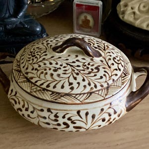 Pode incluir: Uma tigela de cerâmica com tampa, de cor creme, adornada com intrincados padrões florais castanhos. A tigela apresenta duas pegas curvas e uma pequena pega na tampa. O design é simétrico e detalhado.