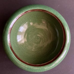 May include: A round, green ceramic bowl with a crackle glaze. The rim is a darker green, and a thin brown band accents the inside edge. The bowl's interior has a textured surface.