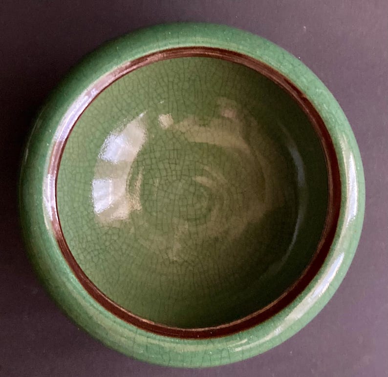 May include: A round, green ceramic bowl with a crackle glaze. The rim is a darker green, and a thin brown band accents the inside edge. The bowl's interior has a textured surface.