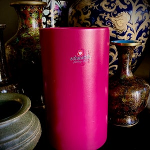 Könnte beinhalten: Eine rosa zylindrische Kerze mit einem weißen Etikett, auf dem "scheurich feeling home" steht. Die Kerze steht auf einem Tisch mit mehreren dekorativen Vasen im Hintergrund.