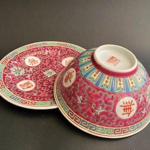 May include: A red and white porcelain bowl and saucer set with Chinese characters. The bowl is upside down, revealing a detailed pattern of blue, yellow, and red accents. The saucer features a matching design with floral and geometric elements.