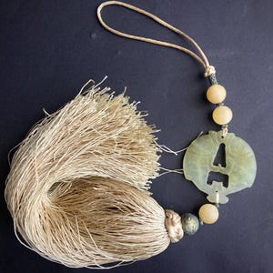 May include: A decorative tassel with a light beige cord and a jade pendant. The pendant features a cut-out design and is accented with light yellow beads. The tassel is made of many strands of light beige thread.