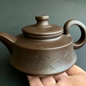 Könnte beinhalten: Braune Keramik-Teekanne mit gebogenem Ausguss, rundem Körper und Schlaufengriff. Der Deckel hat einen kleinen Knopf. Die Teekanne ist mit einem geätzten Blumen- und Vogeldesign verziert. Die Teekanne wird in einer Hand gehalten.