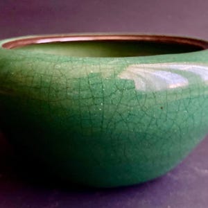 May include: A small, rounded, green ceramic bowl with a crackle glaze. The bowl has a smooth, glossy surface and a dark brown rim. The interior is a darker shade of green. This decorative bowl is suitable for home decor.