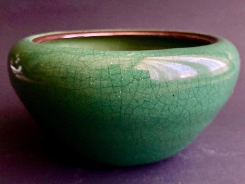 May include: A small, rounded, green ceramic bowl with a crackle glaze. The bowl has a smooth, glossy surface and a dark brown rim. The interior is a darker shade of green. This decorative bowl is suitable for home decor.