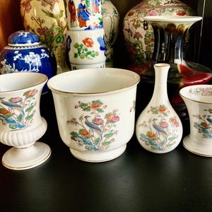 Puede incluir: Un juego de cuatro jarrones de porcelana con un diseño floral y de pájaros. Los jarrones son de varios tamaños y formas, incluyendo un jarrón alto, un cuenco ancho y dos jarrones más pequeños. Todos los jarrones son blancos con ribetes dorados y presentan un patrón floral colorido con pájaros.