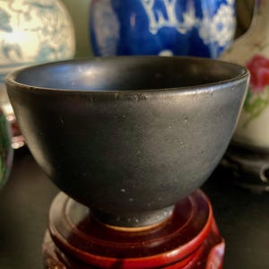 May include: A dark gray ceramic bowl with a rounded shape, set on a small, reddish-brown wooden stand. The bowl has a smooth, matte finish and appears to be handmade. Other decorative ceramic pieces are visible in the background.