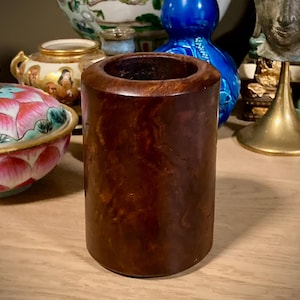 Puede incluir: Un recipiente cilíndrico de madera marrón oscuro con un acabado liso y pulido. El recipiente está vacío y se alza sobre una superficie de color claro. Otros objetos decorativos son visibles en el fondo.