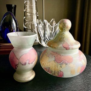 Puede incluir: Dos objetos decorativos de vidrio. Uno es un pequeño jarrón con la parte superior acampanada y base redondeada, con detalles en rosa y morado. El otro es un tarro con tapa con cuerpo bulboso y parte superior redondeada, con detalles similares en rosa y amarillo.