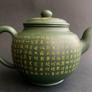 Könnte beinhalten: Dunkelgrüne Keramik-Teekanne mit Deckel und Henkel. Die Teekanne ist mit goldfarbenen chinesischen Schriftzeichen verziert. Ausguss und Henkel sind gebogen. Der Deckel hat einen kleinen Knopf oben. Die Teekanne hat eine klassische, abgerundete Form.