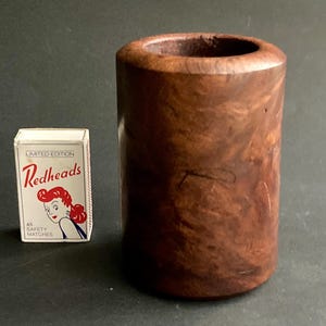 Op de afbeelding: Een cilindrische, donkerbruine houten container met een gladde, gepolijste afwerking. Naast de container staat een doosje "Redheads" lucifers, een limited edition met een vintage design. De houtnerf is zichtbaar, wat bijdraagt aan de natuurlijke uitstraling.