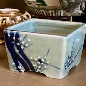 May include: A square ceramic planter with a pale green glaze, featuring hand-painted dark blue branches and small white raised floral accents. The vessel has a wide open top, a smooth finish, and four small feet at the base for stability.