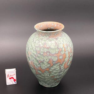 Puede incluir: Un jarrón de cerámica con esmalte cristalino que presenta una base de color naranja terracota superpuesta con patrones de cristales circulares en verde pálido y blanco. Una pequeña caja rectangular de cerillas Redheads está junto al jarrón.