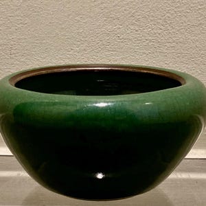 May include: A glossy, green ceramic bowl with a dark interior and a bronze-colored rim. The bowl has a rounded shape and sits on a neutral surface. The green glaze has a crackle effect.