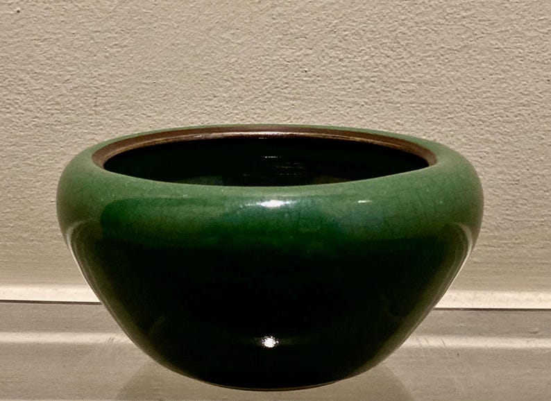 May include: A glossy, green ceramic bowl with a dark interior and a bronze-colored rim. The bowl has a rounded shape and sits on a neutral surface. The green glaze has a crackle effect.