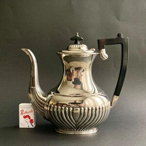 May include: A silver-plated coffee pot with a black handle and a decorative lid. The pot has a curved spout and a ribbed design on the lower half. A small box of "Redheads" matches is next to the pot.