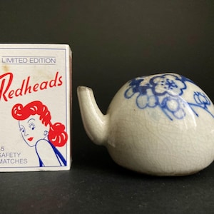 Könnte beinhalten: Eine Vintage-Teekanne aus weißer Keramik mit blauen Blumenmustern und einem kleinen Ausguss. Daneben eine Schachtel "Redheads"-Streichhölzer mit einer Retro-Illustration einer rothaarigen Frau. Auf der Schachtel steht "Limited Edition".