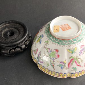 Może przedstawiać: Biała porcelanowa miska z falistym brzegiem, zdobiona kolorowymi motylami i kwiatowymi wzorami. Obok miski znajduje się ciemny drewniany stojak. Miska ma zielono-żółtą obwódkę.