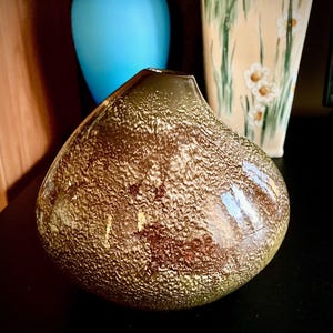 May include: A decorative brown glass vase with a textured surface. The vase has a teardrop shape and a dark brown opening. Other vases are visible in the background.