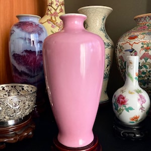 Puede incluir: Una colección de jarrones decorativos de varias formas y colores. El punto focal es un jarrón alto y rosa. Otros jarrones incluyen un jarrón vidriado azul y morado, y un jarrón blanco con diseños florales. Un cuenco plateado descansa sobre un soporte de madera.