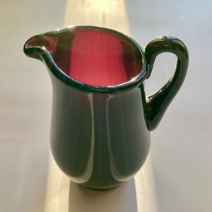 May include: A small, dark green glass creamer with a purple interior and a handle.