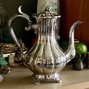 Puede incluir: Una tetera plateada ornamentada, con un pico y asa curvados. La tetera tiene una tapa decorativa y se apoya sobre tres pequeños pies. La superficie de la tetera refleja los objetos circundantes. La tetera es probablemente vintage o antigua.