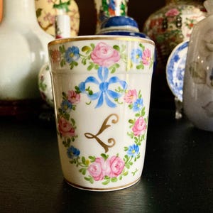 Puede incluir: Una taza de porcelana blanca con un borde dorado y un diseño floral. La taza presenta una cinta azul y la letra "L" en oro.