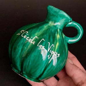 May include: A small, green ceramic jug with a handle and a spout. The jug has a textured surface and white script that reads "Cécile Les Vignes". The jug is a vibrant green colour with yellow accents.