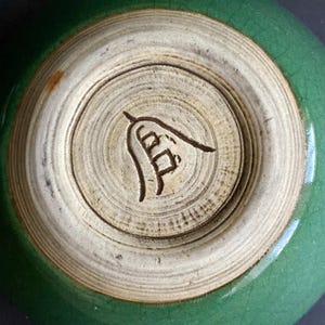 May include: A close-up of the bottom of a green ceramic bowl. The base is a light beige color with a dark brown symbol etched into the center. The bowl's exterior is a glossy, crackled green.