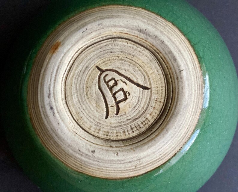May include: A close-up of the bottom of a green ceramic bowl. The base is a light beige color with a dark brown symbol etched into the center. The bowl's exterior is a glossy, crackled green.