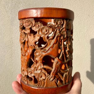 Könnte beinhalten: Ein zylindrischer Pinselhalter aus braunem Holz mit aufwendigen Durchbruchschnitzereien aus wirbelnden Wolken und organischen Formen. Das Gefäß hat einen glatten, massiven Rand an der Ober- und Unterseite sowie einen hohlen Innenraum.