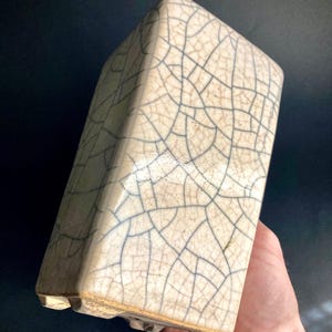 Może przedstawiać: Biała ceramiczna doniczka z pękniętą glazurą. Doniczka ma kwadratowy kształt z zaokrąglonymi krawędziami i lekko uniesioną podstawą.