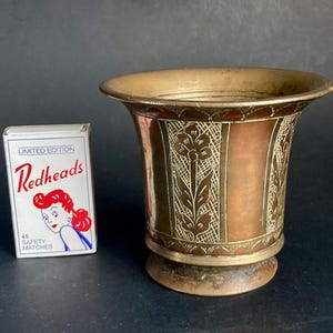 Puede incluir: Un jarrón vintage de latón y cobre con diseños florales grabados. El jarrón tiene una parte superior acampanada y una base redonda. Una caja de cerillas "Redheads" está al lado del jarrón.