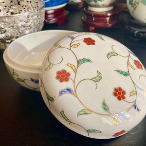 May include: A white ceramic trinket box with a lid, decorated with a floral pattern of red flowers, green leaves, and gold accents. The box is open, revealing a plain white interior. The design is reminiscent of Asian porcelain.