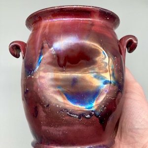 Op de afbeelding: Een keramische vaas met een glanzende, iriserende afwerking in tinten diep rood, koper en metallic blauw. De vaas heeft een afgerond lichaam, een brede opening en twee kleine, gebogen handvatten aan weerszijden van het object.