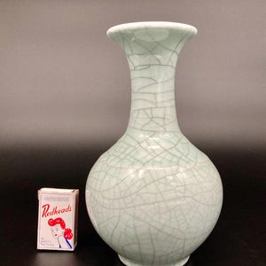 May include: A pale celadon green ceramic vase with a bulbous body and a flared rim, featuring a prominent dark crackle glaze pattern throughout the surface. A small rectangular box of Redheads safety matches is positioned next to the vase.