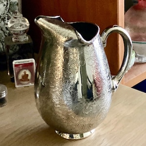 May include: A silver-colored pitcher with an ornate floral pattern. The pitcher has a curved handle and a spout for pouring. The surface reflects light, creating highlights and shadows. The pitcher is likely made of metal and is a decorative item.