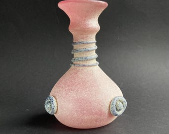 May include: A pink glass vase with a textured surface. The vase has a bulbous body, a narrow neck with a spiral design, and a flared top. Two blue and white circular accents are on the sides.